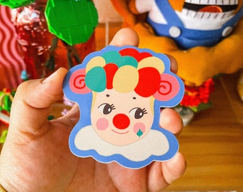 May include: A colorful die-cut sticker featuring a cartoon clown face with a red nose, a diamond-shaped tear, and a blue outline. The clown has a white collar and a multi-colored hairpiece. The sticker is held in a hand.