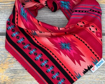 May include: A vibrant dog bandana in shades of red and pink, showcasing a southwestern-inspired pattern. The design incorporates black, turquoise, and pink geometric elements and stripes. The bandana is knotted at the top and includes a small tag with the word "Puppers".