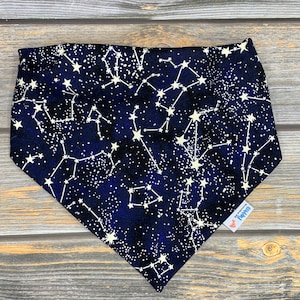 May include: A navy blue bandana with a white constellation pattern and white stars. The bandana has a white tag with the text "Pawsitive Pups" on it.
