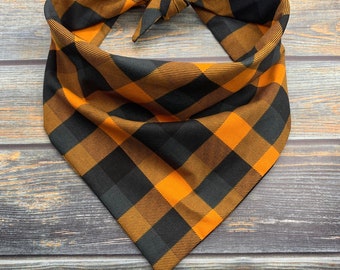May include: A plaid dog bandana in orange and black. The bandana is tied at the top and has a metal ring. The fabric has a classic buffalo check pattern. The bandana is displayed on a wooden surface.