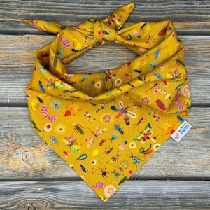 May include: A yellow bandana with a pattern of colorful insects and flowers. The bandana is tied in a knot at the top and has a small tag that says "The Pawsitive."