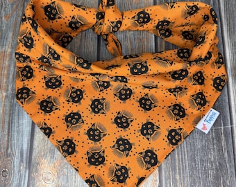 Peut inclure: Bandana orange pour animaux de compagnie avec un motif répété de têtes de chats noirs et d'étoiles. Le bandana est noué en haut et porte une petite étiquette blanche avec un logo. Le tissu est doux, idéal pour Halloween ou au quotidien.