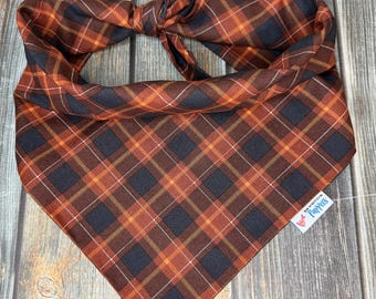 May include: A plaid dog bandana in shades of orange, brown, and navy blue. The bandana is tied at the top and features a repeating square pattern. A small tag with text is visible on the bottom right.