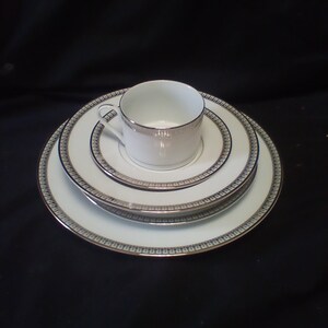 May include: A white porcelain teacup and saucer set with silver trim, stacked on a matching plate set. The plates and cup have a delicate blue and silver patterned border. The set is displayed on a black surface.