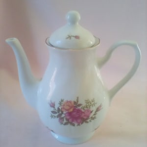 May include: White porcelain teapot with a floral design featuring pink, yellow, and green roses. The teapot has a lid and a gold trim around the rim.