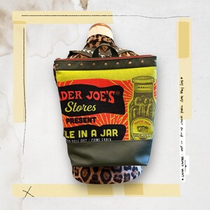 May include: A unique backpack featuring a design inspired by Trader Joe's. The bag has a yellow and red front panel with the text "Trader Joe's Stores Present Pickle in a Jar". The bag has a leopard print strap and bottom.