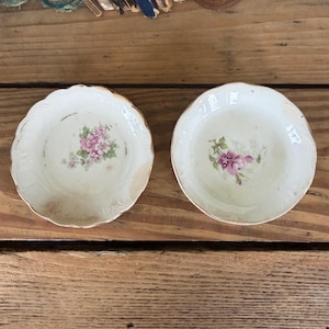 May include: Two antique, off-white porcelain saucers with floral designs. Each saucer features a delicate pink and green floral pattern in the center and a gold trim around the edge. The saucers are placed on a wooden surface.