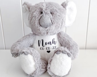 May include: Gray plush koala bear with white fur on its paws and ears. The bear is wearing a white shirt with the name "Noah" and the date "01.01.22" printed in black.