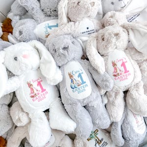May include: A collection of plush bunny rabbits with personalised embroidered patches. Each bunny has a different name and the text "1st Easter 2025". The bunnies are in various colours, including white, grey, and brown.