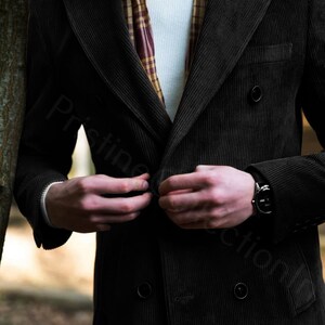 May include: A black corduroy double-breasted jacket with a button closure. The jacket has a pocket on the left side and a brown and white plaid scarf is worn underneath.
