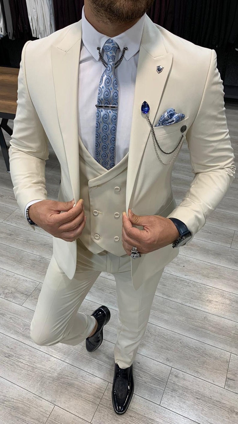 May include: A beige suit with a vest and a blue patterned tie. The suit has a double-breasted vest and a chain detail on the jacket.