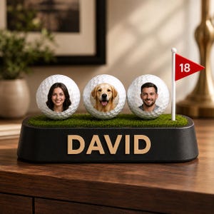 May include: A black display base with the name DAVID in gold letters, featuring three white golf balls with printed portraits of a woman, a golden retriever, and a man, plus a small white flagpole with a red flag displaying the number 18.