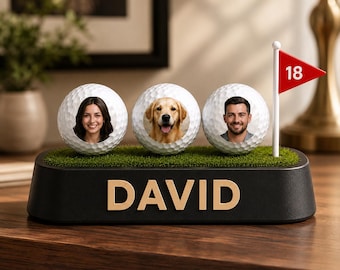 May include: A black display base with the name DAVID in gold letters, featuring three white golf balls with printed portraits of a woman, a golden retriever, and a man, plus a small white flagpole with a red flag displaying the number 18.