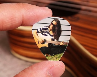 May include: A clear guitar pick with a photo of a person playing a guitar. The person is wearing a striped shirt and has long dark hair. The photo is set against a white background with black lines.
