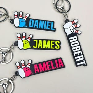 Könnte beinhalten: Schlüsselanhänger mit Bowlingkegel- und Kugeldesigns in Blau, Grün und Pink, jeweils mit einem Namen: Daniel, James, Robert und Amelia. Die Schlüsselanhänger haben silberfarbene Metallringe.