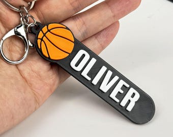 May include: A black keychain with the name "OLIVER" in white lettering. The keychain features an orange and black basketball design. The keychain is attached to a silver keyring.