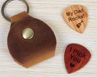 May include: Two wooden guitar picks with engraved messages. One pick is light brown with the text "My Dad Rocks!" and a small guitar icon. The other pick is dark red with the text "I Pick You" and a small heart icon. A brown leather guitar pick holder with a metal ring is also included.