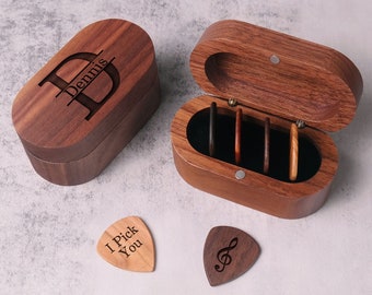 May include: A wooden box with a personalized initial and the name "Dennis" engraved on the lid. The box is open and contains a set of six guitar picks. Two additional guitar picks are shown on a light wood surface. One pick has the text "I Pick You" engraved on it, and the other has a treble clef symbol.