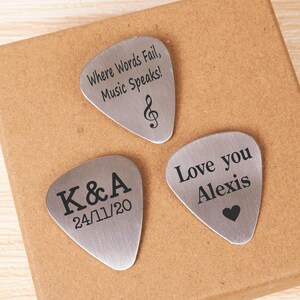 May include: Three silver guitar picks with engraved messages. The first pick says "Where Words Fail, Music Speaks!" with a treble clef symbol. The second pick says "K&A 24/11/20". The third pick says "Love you Alexis" with a heart symbol.