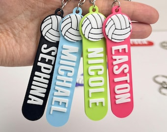 May include: Four personalized volleyball keychains in black, light blue, green, and pink. Each keychain features a white and black volleyball design and a name in white block letters: Sephina, Michael, Nicole, and Easton. The keychains are attached to silver keyrings.