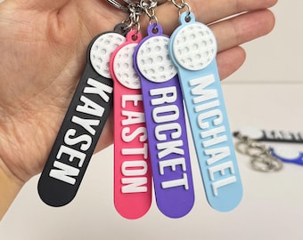 May include: Four colorful keychains with golf ball accents. Each keychain has a different color: black, pink, purple, and light blue. Each keychain has a name in white lettering: KAYSEN, EASTON, ROCKET, and MICHAEL. The keychains are attached to silver keyrings.