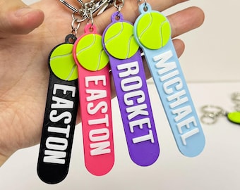 May include: A collection of personalized tennis-themed keychains. Each keychain features a tennis ball design and a name in white lettering on a colored background. Colors include black, pink, purple, and light blue. The keychains are attached to silver key rings.