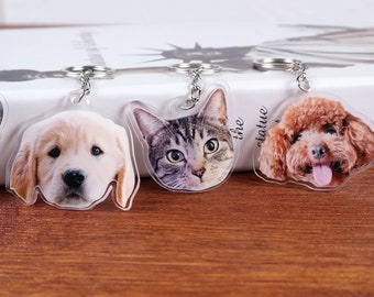 May include: Three keychains featuring clear plastic charms with images of a golden retriever puppy, a tabby cat, and a brown poodle puppy. The keychains are on a wooden surface.