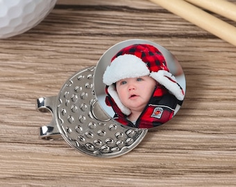 May include: A silver golf ball marker with a round photo insert featuring a baby wearing a red and black plaid hat. The photo insert is attached to the marker with a magnet.
