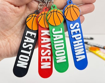 May include: Four personalized basketball keychains in black, red, green, and blue. Each keychain features a basketball design and a name in white lettering: Easton, Kaysen, Jaidon, and Sephina. The keychains are attached to silver keyrings.