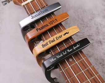 May include: Four guitar capo clamps with different engraved messages: "Jonathon", "Psalms 101:1", "Best Dad Ever" with a mustache symbol, and "Where Words Fail, Music Speaks!"
