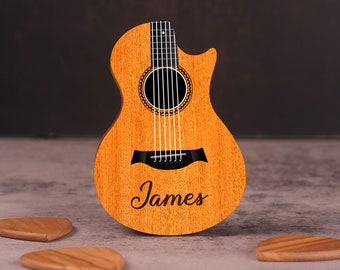 May include: A wooden acoustic guitar with a natural finish. The guitar has a black pickguard and a black name inscription that reads "James". The guitar is surrounded by three wooden guitar picks.