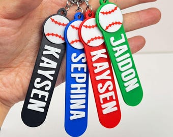 May include: Four baseball-themed keychains in black, blue, red, and green. Each keychain features a baseball design and a name in white block letters. The keychains are attached to a silver keyring.