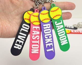 May include: Keychains with softball designs in black, pink, purple, and green. Each keychain has a yellow softball with red stitching and a name in white lettering: Oliver, Easton, Rocket, and Jaidon. The keychains are attached to silver keyrings.