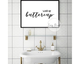 May include: A black and white bathroom sign with the text "Wash up buttercup" in a cursive font. The sign is hanging on a white tiled wall above a white porcelain sink with gold fixtures.