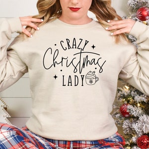 Może przedstawiać: Beżowa bluza z napisem "Crazy Christmas Lady" czarnymi literami. Obok tekstu znajduje się mały obrazek kubka z gorącą czekoladą z bitą śmietaną i płatkiem śniegu.