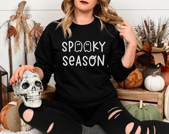 May include: Black sweatshirt with the text "Spooky Season" and ghost graphics. The image includes a decorative skull, pumpkins, and fall foliage, creating a Halloween theme.