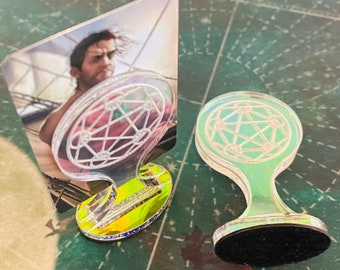 Pode incluir: Duas peças de jogo transparentes e iridescentes com um design em forma de estrela. Uma peça tem a imagem de um homem num navio. A outra peça tem uma base circular preta. Ambas as peças têm uma pega curva.