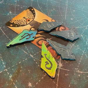 May include: Five iridescent, triangular game pieces with different symbols on each. The pieces are on top of a card with a dark background and a stylised tree with a star at the top.