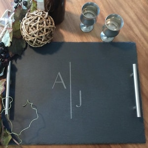 May include: A black slate serving tray with silver handles and the initials "AJ" etched in the center. The tray is on a wooden surface with a decorative ball and two silver goblets.