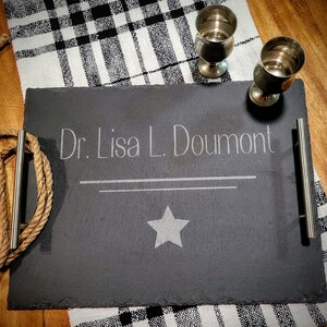 May include: A black slate serving tray with silver handles. The tray is engraved with the text "Dr. Lisa L. Doumont" and a star.