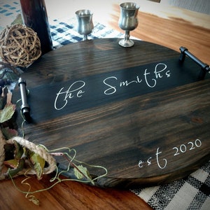 May include: A round wooden serving tray with black stain and two metal handles. The tray has a black stripe across the center with the text "the Smiths est 2020" written in white.