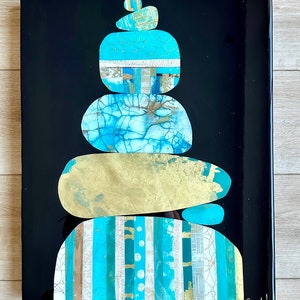 Puede incluir: Una pila de cinco piedras pintadas de forma abstracta en tonos azules, verdes y dorados, sobre un fondo negro. Las piedras est&aacute;n dispuestas en una pila zen, con la piedra m&aacute;s peque&ntilde;a en la parte superior. La firma del artista es visible en la esquina inferior derecha.
