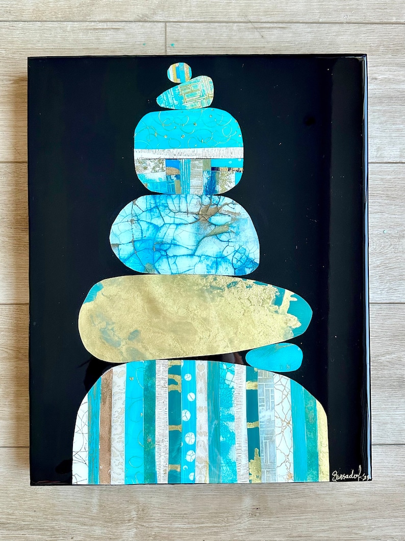 Puede incluir: Una pila de cinco piedras pintadas de forma abstracta en tonos azules, verdes y dorados, sobre un fondo negro. Las piedras est&aacute;n dispuestas en una pila zen, con la piedra m&aacute;s peque&ntilde;a en la parte superior. La firma del artista es visible en la esquina inferior derecha.