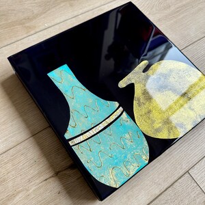 Puede incluir: Un lienzo cuadrado negro con dos jarrones de cer&aacute;mica abstractos. Un jarr&oacute;n es azul turquesa con detalles dorados y el otro es dorado con interior blanco.