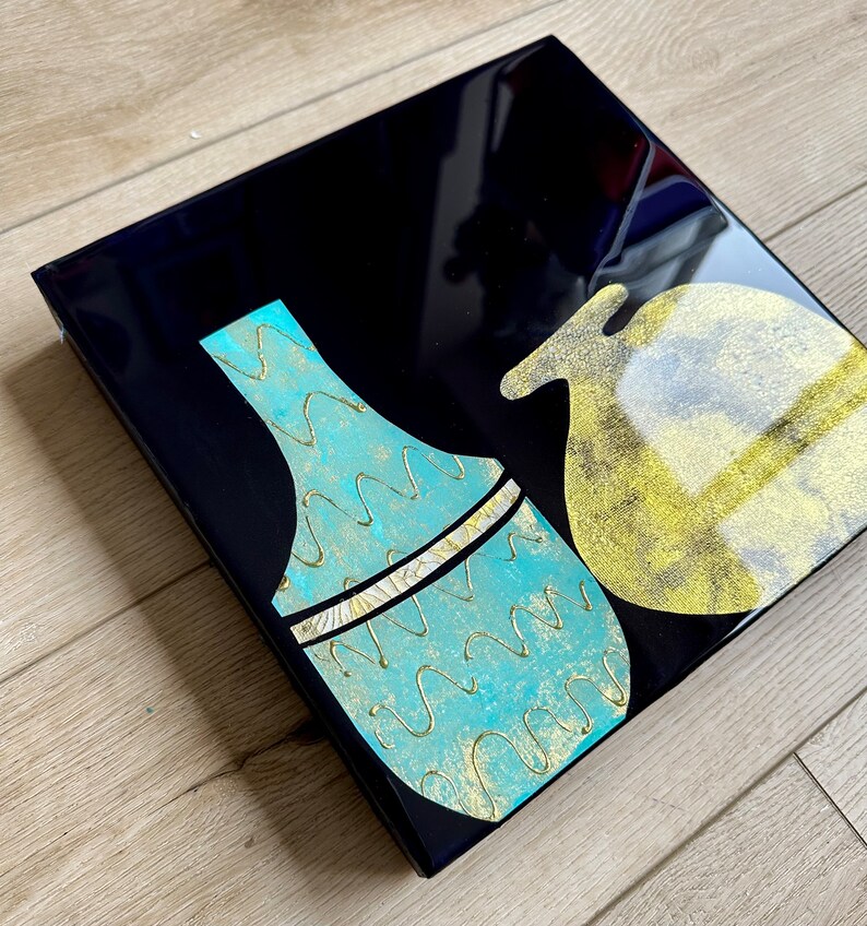 Puede incluir: Un lienzo cuadrado negro con dos jarrones de cer&aacute;mica abstractos. Un jarr&oacute;n es azul turquesa con detalles dorados y el otro es dorado con interior blanco.