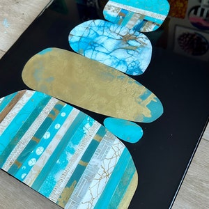 Puede incluir: Pintura abstracta de piedras apiladas en tonos de azul, dorado y blanco sobre un fondo negro.