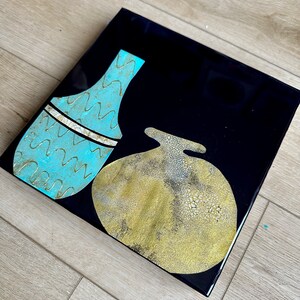 Puede incluir: Dos jarrones de cer&aacute;mica abstractos sobre un fondo negro. Un jarr&oacute;n es de color azul claro con un patr&oacute;n ondulado y un acento dorado. El otro jarr&oacute;n es de color dorado con una superficie texturizada.