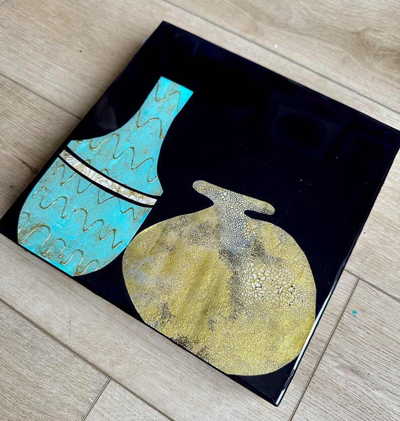 Puede incluir: Dos jarrones de cer&aacute;mica abstractos sobre un fondo negro. Un jarr&oacute;n es de color azul claro con un patr&oacute;n ondulado y un acento dorado. El otro jarr&oacute;n es de color dorado con una superficie texturizada.