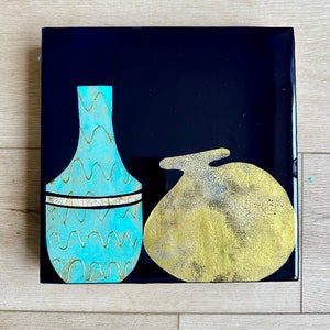 Puede incluir: Un jarr&oacute;n de cer&aacute;mica azul y dorado con un patr&oacute;n ondulado y una banda dorada se encuentra junto a un objeto de cer&aacute;mica dorado y blanco sobre un fondo negro.