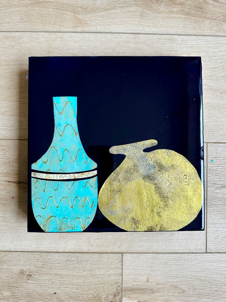 Puede incluir: Un jarr&oacute;n de cer&aacute;mica azul y dorado con un patr&oacute;n ondulado y una banda dorada se encuentra junto a un objeto de cer&aacute;mica dorado y blanco sobre un fondo negro.